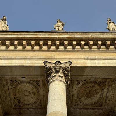 Drie beelden boven een klassieke zuilengalerij met gedetailleerde ornamenten en een blauwe lucht.