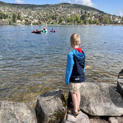 Mats staat aan de oever van een meer, waarop meerdere bootjes varen en op de achtergrond de bergen rondom het meer