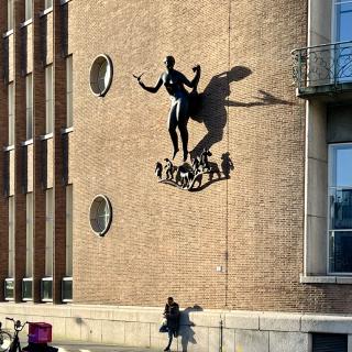 Een man staat tegen een monumentaal gebouw geleund, terwijl de schaduwen over het gebouw trekken