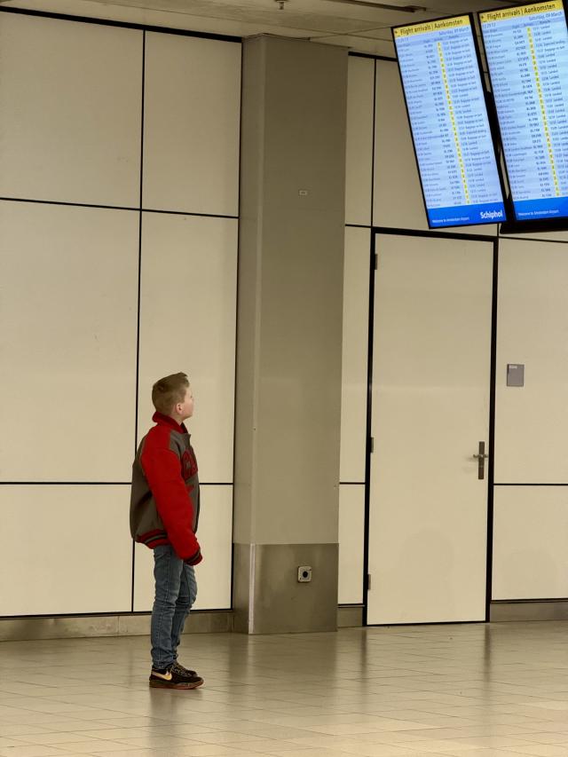 Een jongen in een rode en grijze jas kijkt naar aankomstborden in een luchthavenhal.
