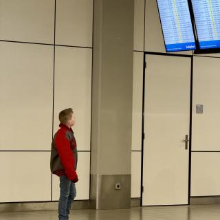 Een jongen in een rode en grijze jas kijkt naar aankomstborden in een luchthavenhal.