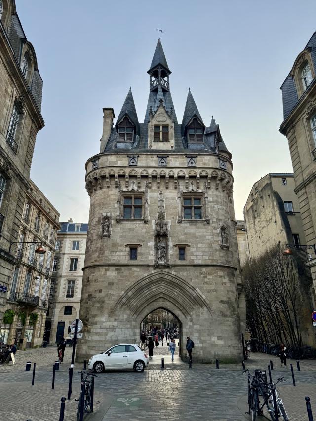 Middeleeuwse poort met torens in Bordeaux, omringd door gebouwen en mensen. Een witte auto staat ervoor op een geplaveide straat.
