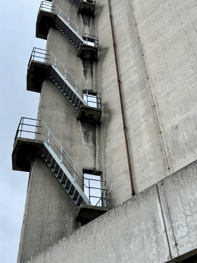 Een betonnen toren met een zigzag trap en metalen railingen, onder een grijze hemel.