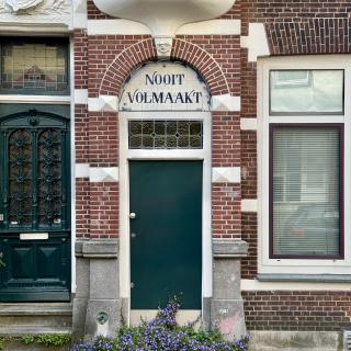 Rode bakstenen gevel met groene deur en decoratieve boog. Boven de deur staat NOOIT VOLMAAKT. Een buste en paarse bloemen voegen charme toe.
