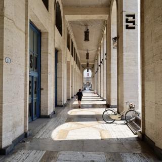 Een lange, overdekte gang met bogen en blauwe deuren, verlicht door hanglampen. Een persoon loopt naar voren en een fiets staat tegen de muur.