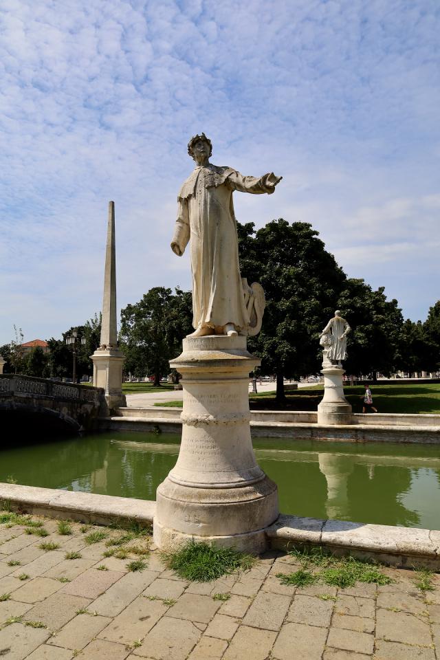 Standbeeld van een man op een sokkel bij een vijver met een obelisk en bomen in de achtergrond.