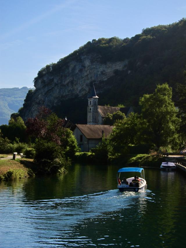 Een bootje vaart over een kalme rivier langs een schilderachtig dorpje met een kerkje en een steile rotswand, omgeven door groen en bomen onder een heldere blauwe lucht.