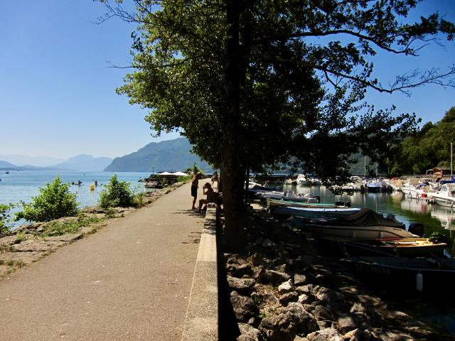 Een pad langs een meer met boten, mensen die ontspannen en bergen in de verte onder een heldere blauwe lucht.