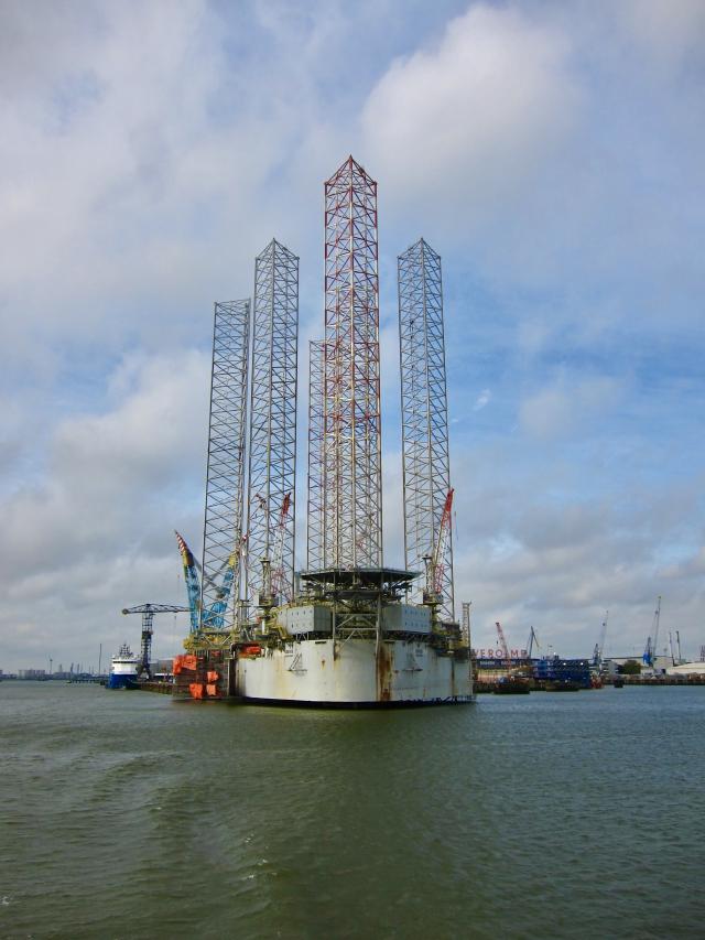 Een offshore booreiland met vier hoge torens in een haven, omgeven door water en kranen onder een bewolkte hemel.