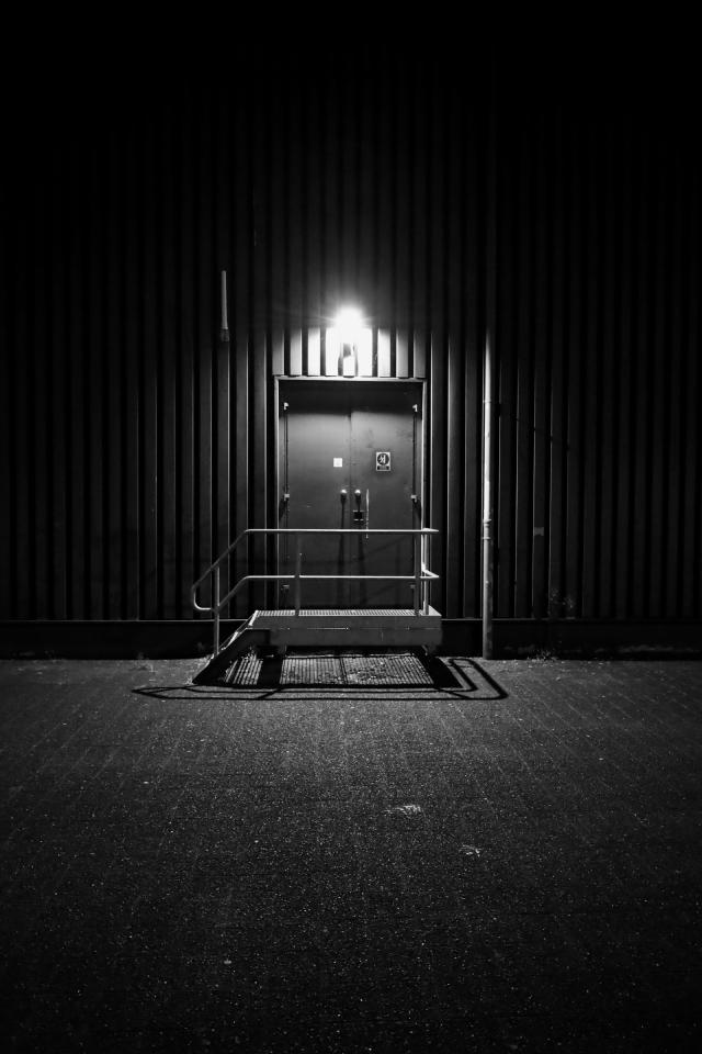 Donkere nacht met een verlichte deur in een metalen wand, omgeven door een leeg asfaltplein.